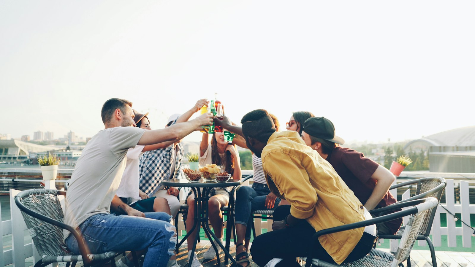 Friends seated around a rooftop table clinking glasses and bottles in a toast